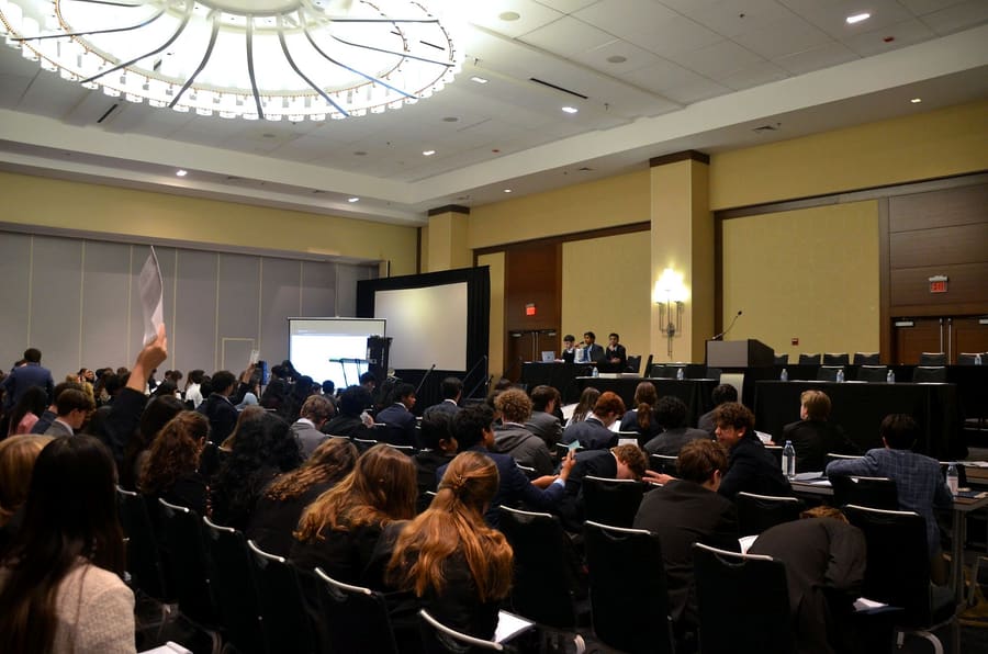 A packed ballroom of delegates in committee, a placard raised to be recognized by the chair